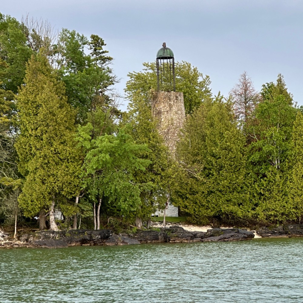 Stone tower with a metal roof surrounded by trees near a shoreline.