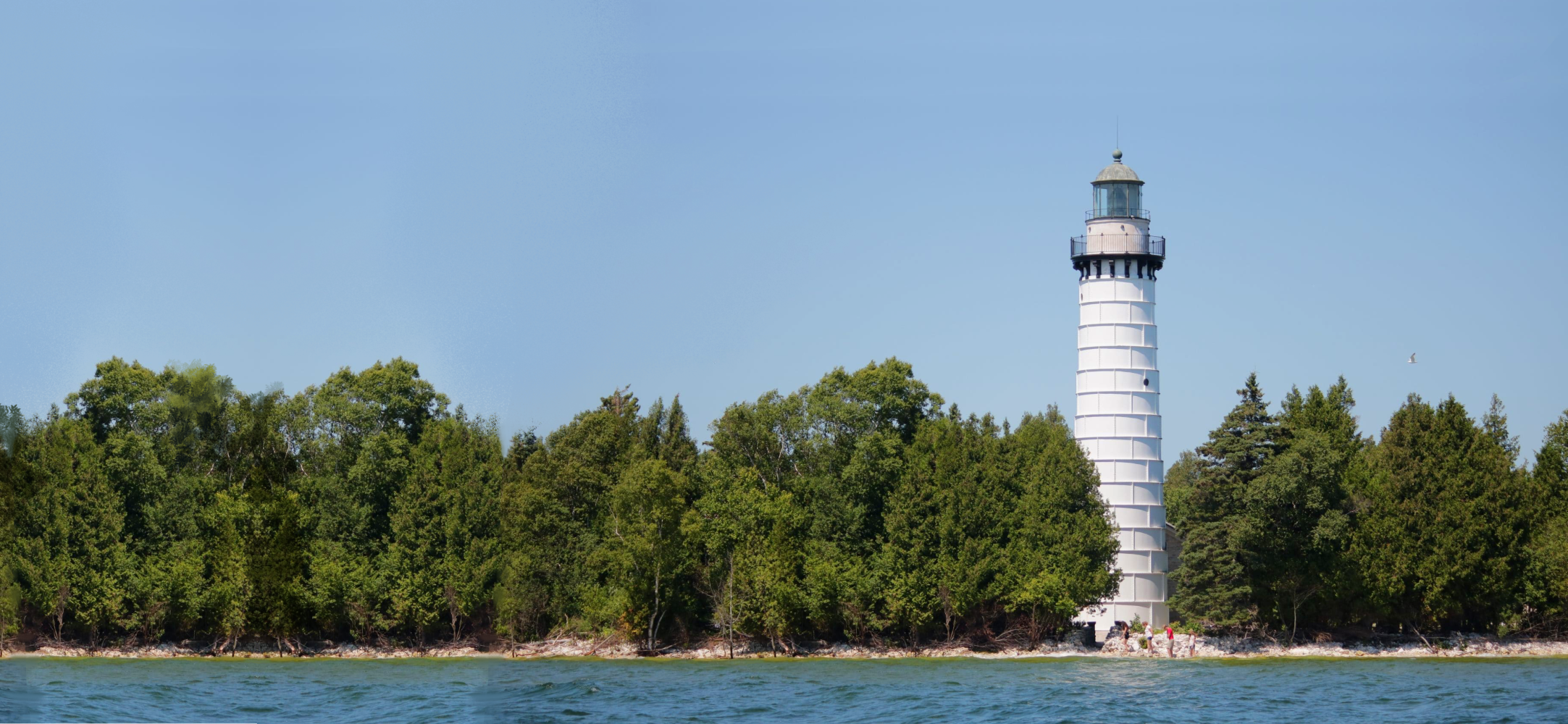 White lighthouse surrounded by trees on a shoreline with blue sky.