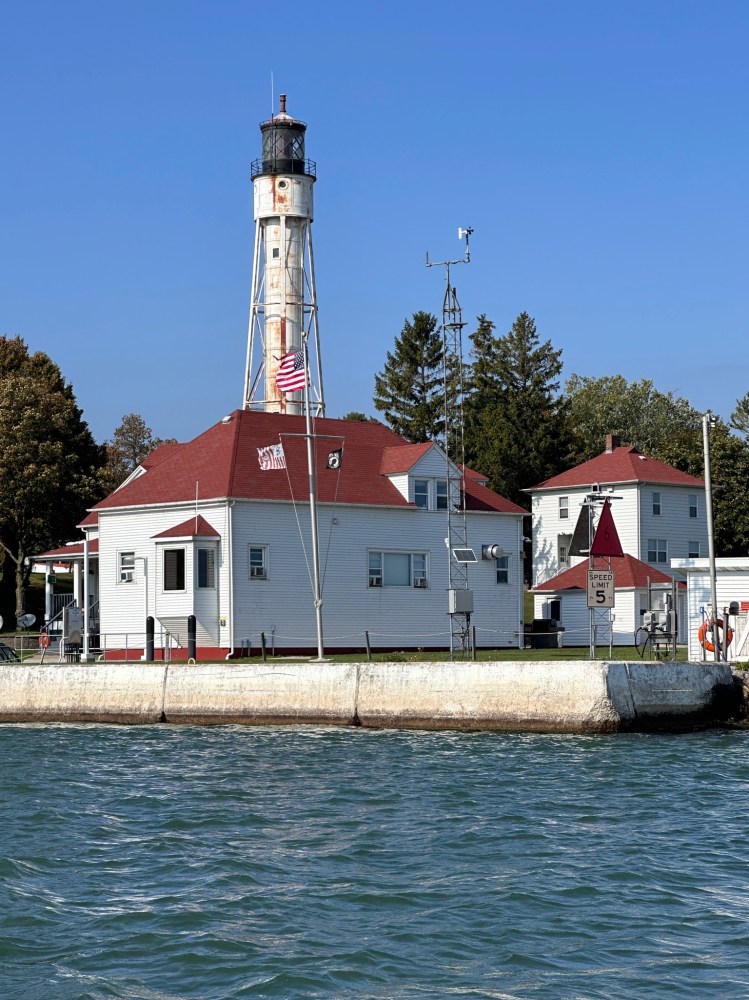 Tall lighthouse with red-roofed building, flagpole, near water under blue sky.