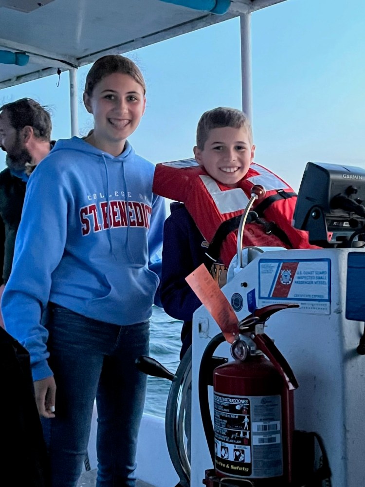 Two kids on a boat, one wearing a life jacket, smiling near a fire extinguisher.
