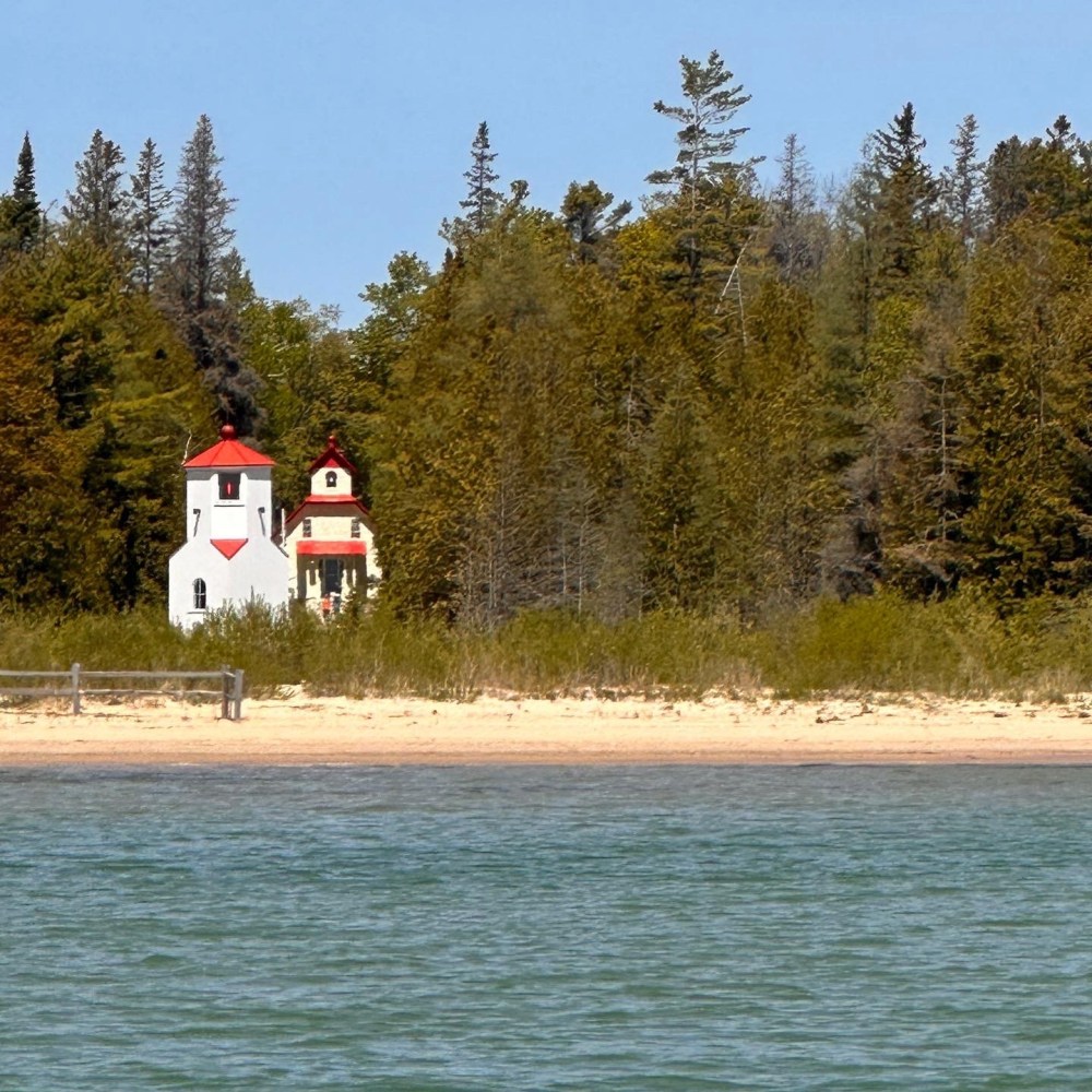 Lighthouse with red roof among trees near sandy shore and blue water.