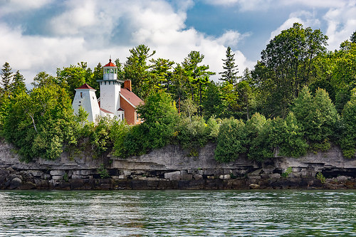 A lighthouse surrounded by trees near a rocky shoreline with calm water.