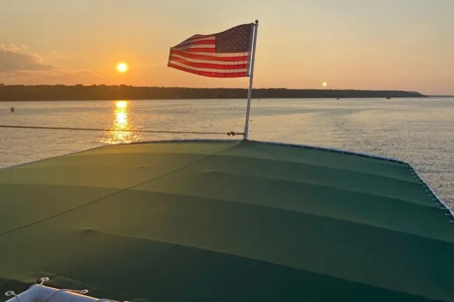 Boat with USA flag at sunset on calm water, green roof in foreground.
