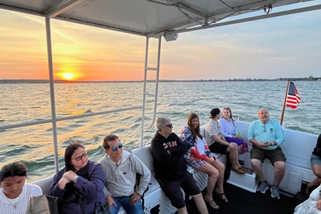 People seated on a boat at sunset, with a U.S. flag and lake in the background.
