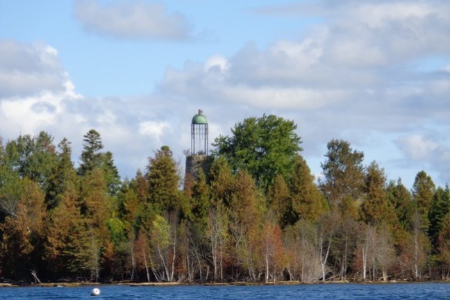 Lighthouse tower behind colorful trees by a lake under a cloudy sky.