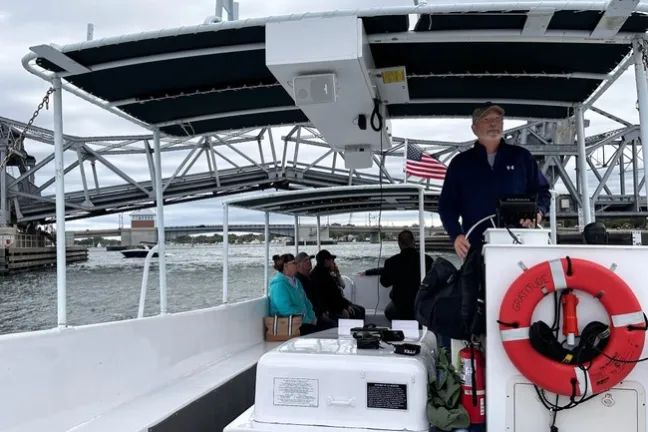 People on a boat under a bridge, with a man steering and a lifebuoy visible.