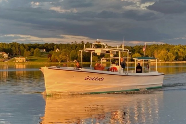 A small boat named Gratitude on a lake during sunset, with people onboard.
