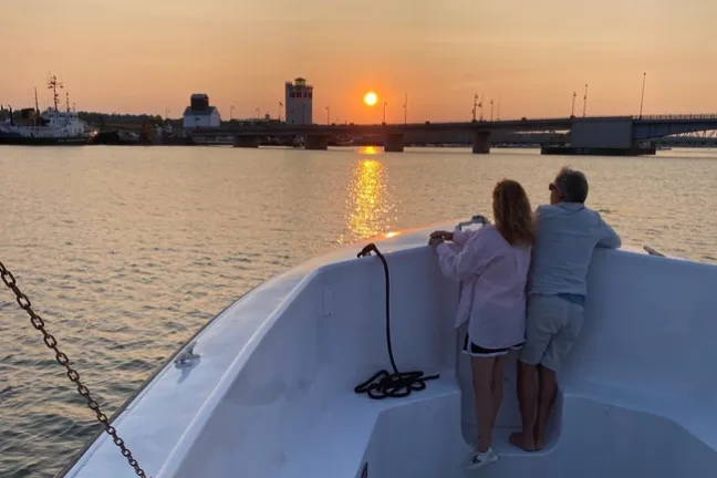 Two people on a boat deck watching the sunset over calm water and a distant bridge.